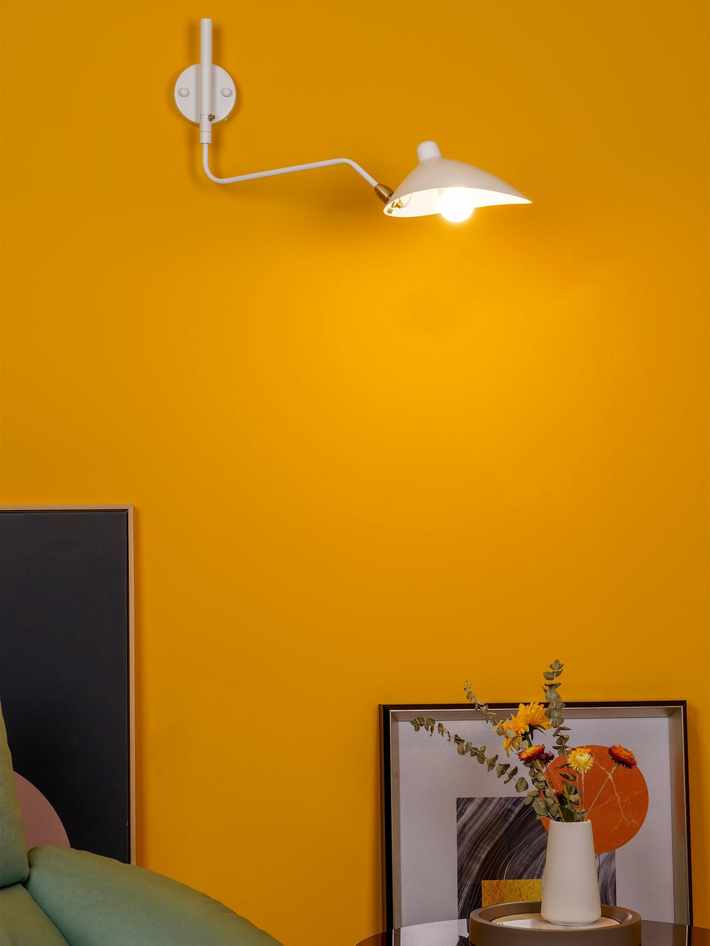 White Clérachic wall light with a curved arm and a lit cone head, on a bright mustard wall above a framed print.