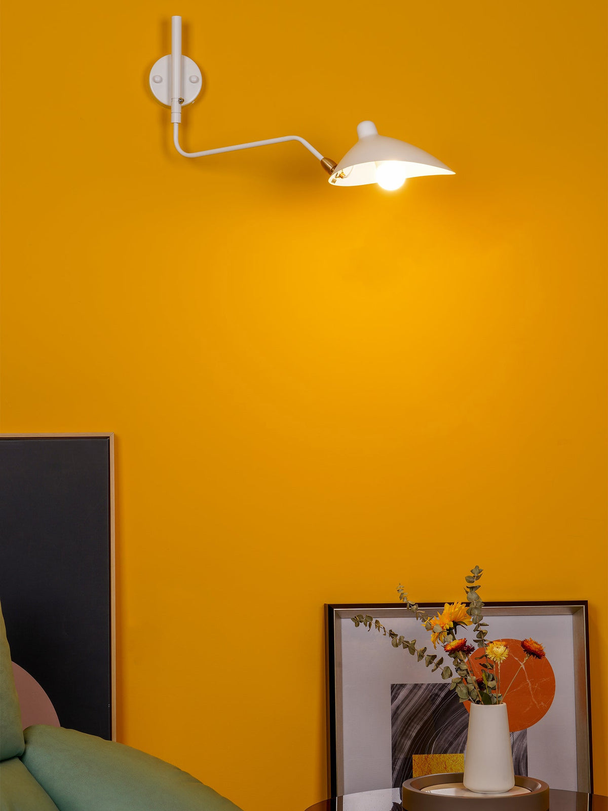 White Clérachic wall light with a curved arm and a lit cone head, on a bright mustard wall above a framed print.