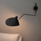 Close view of the black cone shade and brass joint on a Clérachic wall light, lit against a grey wall above a linen cushion.
