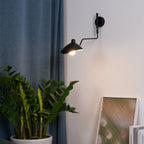 Clérachic black wall light lit on a pale grey wall, with a blue curtain at the left and a large green plant below.