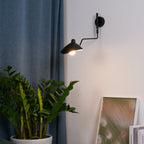 Clérachic black wall light with a cone shade and a curved arm, on a pale wall beside a blue curtain and a green plant.