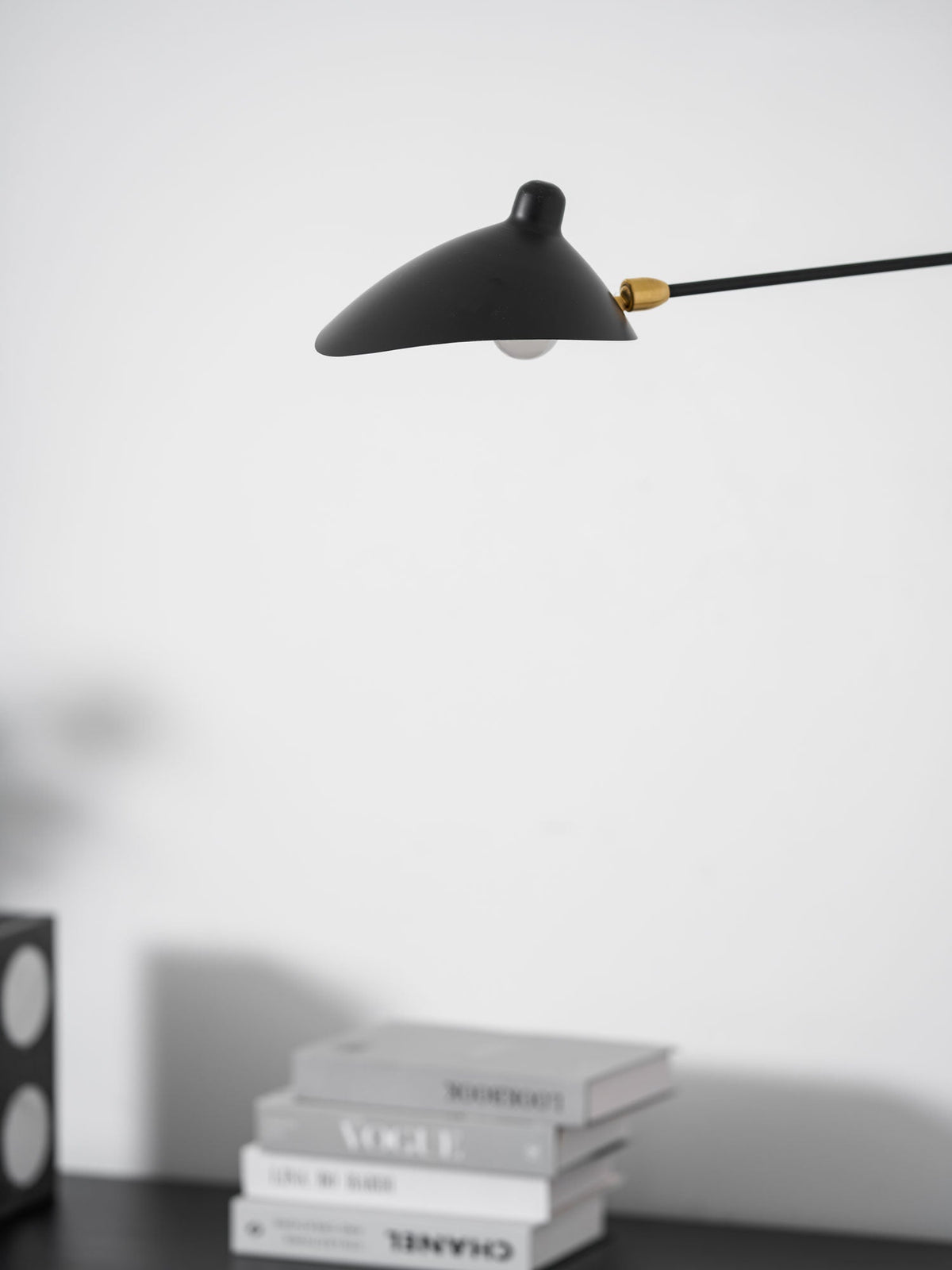 Close view of the black cone shade and brass joint on this Clérachic wall light, with books on a black chest below.