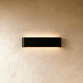 Clérachic slim black aluminium wall light on a warm limewash wall, glowing softly above and below its rounded ends.