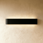 Clérachic long black aluminium wall light lit, washing a warm glow up and down across a textured limewash wall.