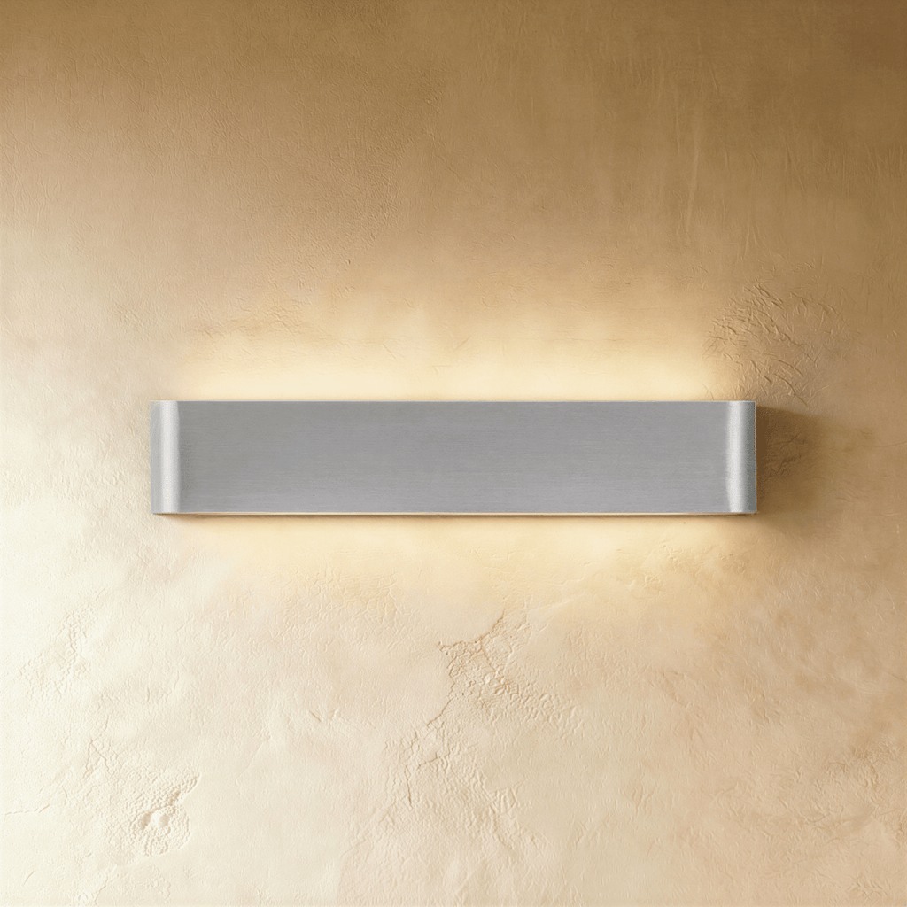 Clérachic longer brushed silver wall light with curved ends, spreading warm light up and down a golden plaster wall.