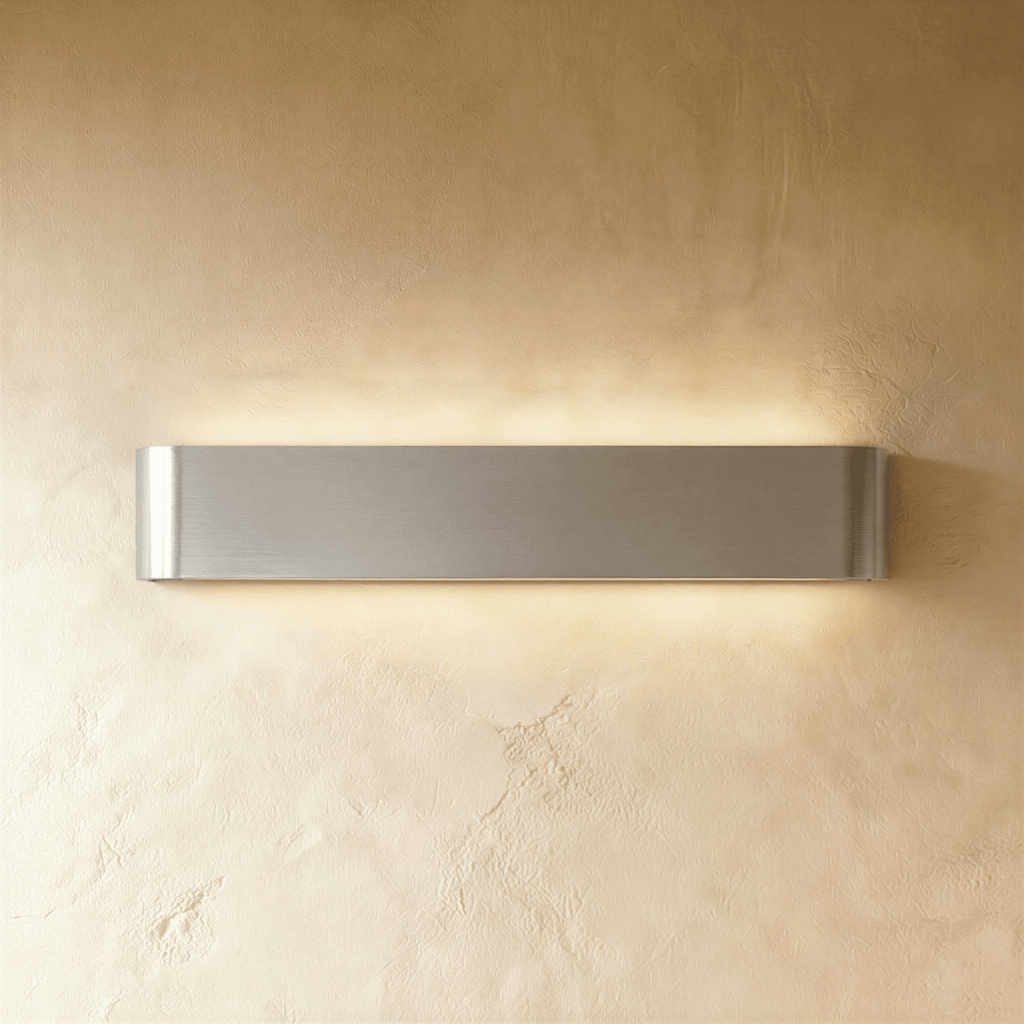 Clérachic long brushed silver aluminium wall light lit, its curved end caps and metal grain clear on a warm wall.