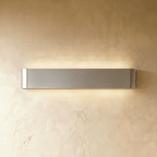 Clérachic long brushed silver aluminium wall light lit, its curved end caps and metal grain clear on a warm wall.