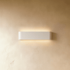 Clérachic slim white aluminium wall light with rounded ends, casting a soft warm glow above and below on cream plaster.