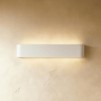 Clérachic longer white aluminium wall light with rounded ends, glowing warmly above and below on a plaster wall.