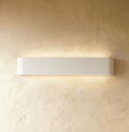Clérachic long white aluminium wall light lit, a wide band of warm light spreading over golden textured plaster.