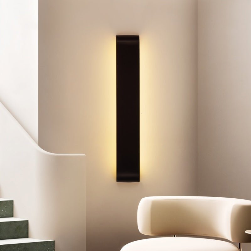 Clérachic tall black aluminium wall light mounted vertically beside a curved stair, lighting the wall behind an armchair.