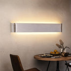Clérachic long brushed silver aluminium wall light glowing above a small round dining table with fruit and a vase.