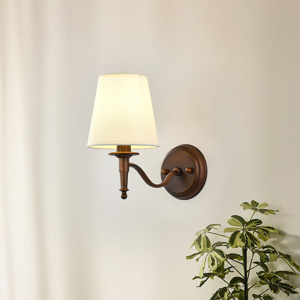 Clérachic wall light with a curved S-shaped arm and a white tapered shade, glowing softly on a cream wall beside a leafy plant.