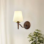 Clérachic wall light with a curved S-shaped arm and a white tapered shade, glowing softly on a cream wall beside a leafy plant.