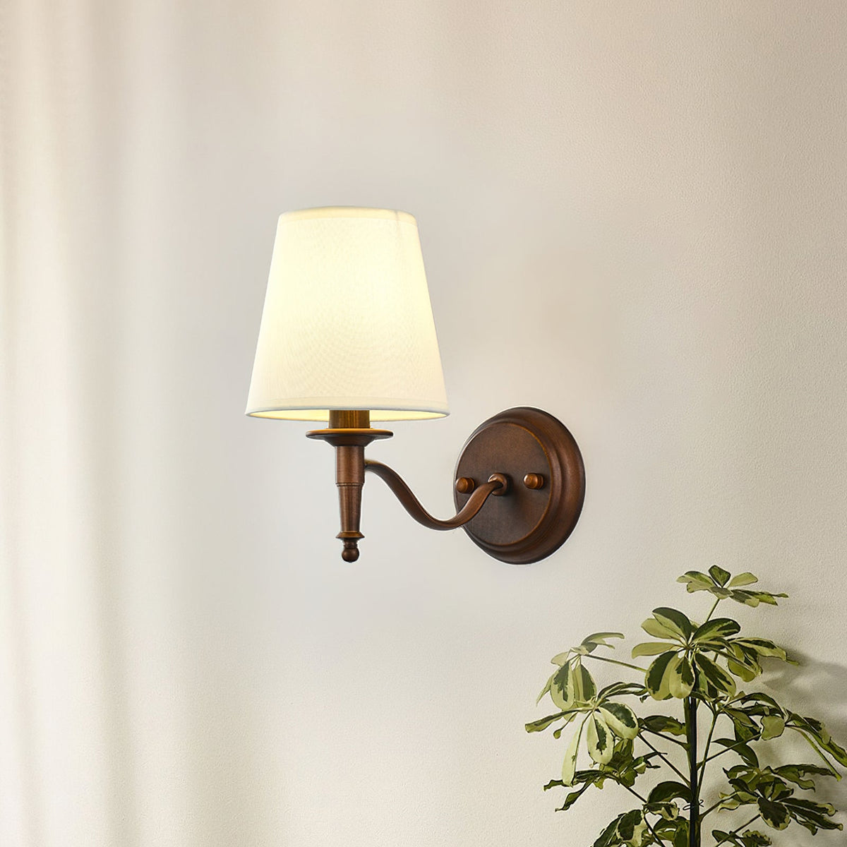Clérachic wall light with a curved S-shaped arm and a white tapered shade, glowing softly on a cream wall beside a leafy plant.