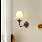 Curved-arm Clérachic wall light on a warm cream wall, next to a sheer curtain and a small gold oval frame.
