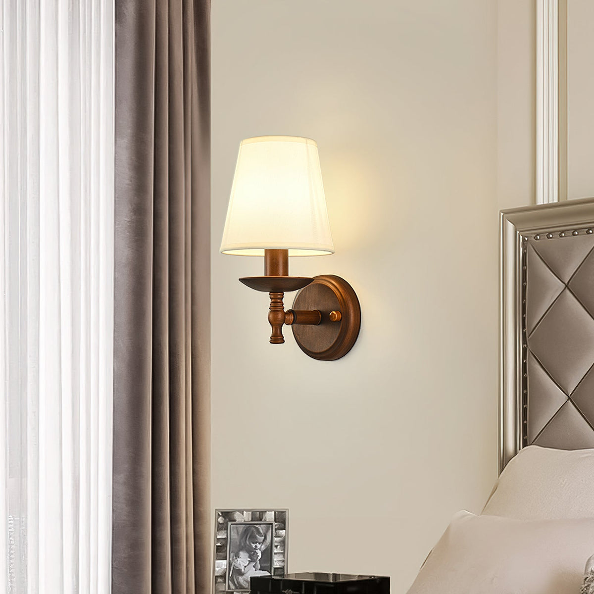 Clérachic wall light with a straight arm and glowing white shade, between a mauve curtain and a padded headboard in a bedroom.