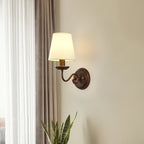 Antique brown Clérachic wall light with a swan-neck arm, lit on a cream wall between a sheer curtain and a taupe drape.