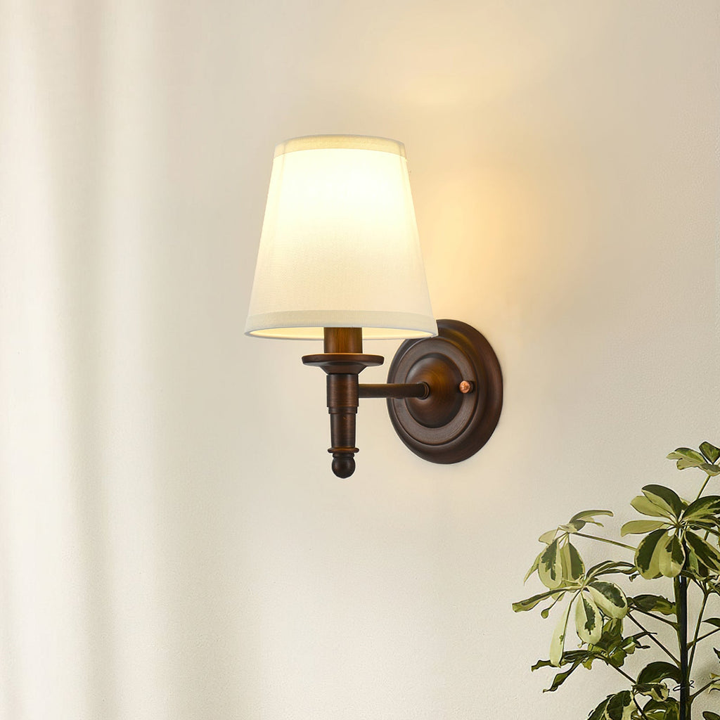 Close view of the straight-arm Clérachic wall light lit warm on a cream wall, with the leaves of a plant at the lower right.