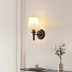 Small straight-arm Clérachic wall light on a pale wall above a dark side table, with a vase of flowers at the right.