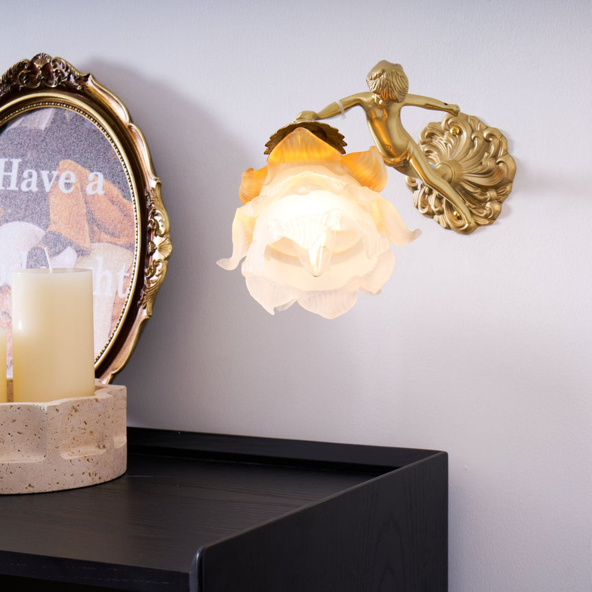 A Clérachic cherub wall light with a frosted glass rose shade glowing beside a candle and a framed print above a black cabinet.