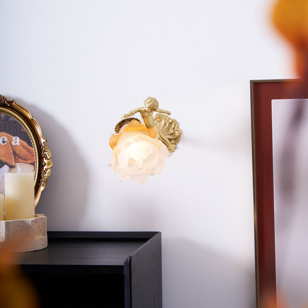 Small in the frame, this Clérachic cherub wall light with its rose-shaped glass shade sits on a pale wall beside a candle and a dark picture frame.