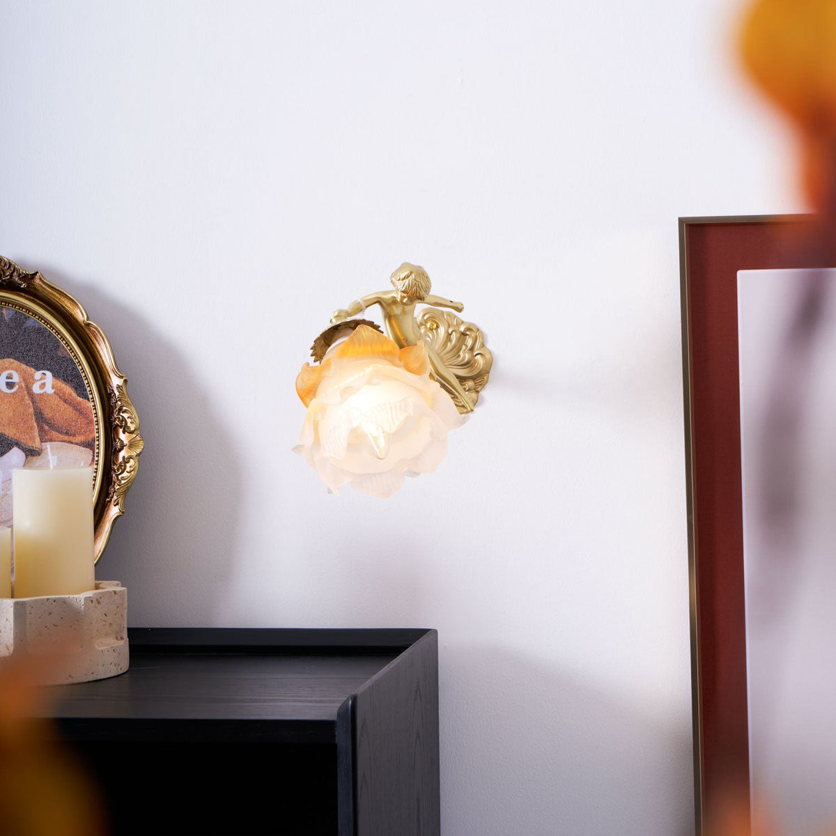Small in the frame, this Clérachic cherub wall light with its rose-shaped glass shade sits on a pale wall beside a candle and a dark picture frame.