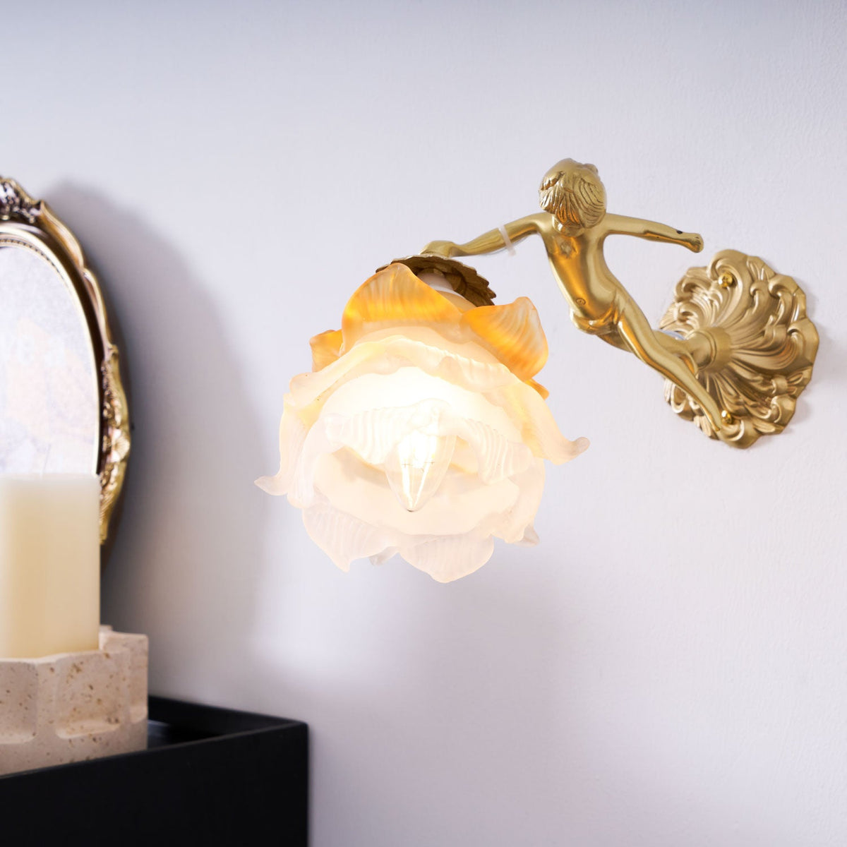 Closer view of the Clérachic cherub wall light, its frosted rose shade lit warm above a candle on a black cabinet.