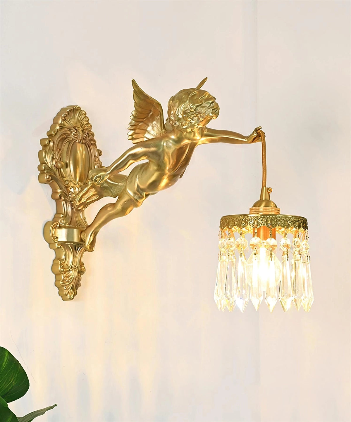 The cherub on this Clérachic wall light reaches out to a crystal-fringed shade, lit warm against a cream wall with a green leaf below.