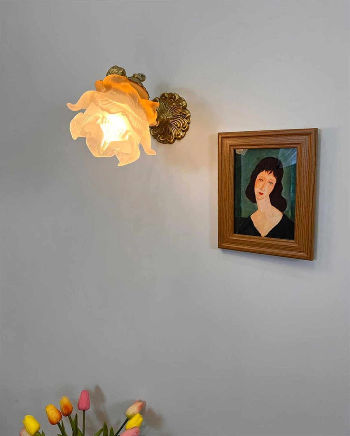 Lit rose-shaped shade of a Clérachic wall light on a grey wall, next to a framed portrait with tulips at the bottom edge.