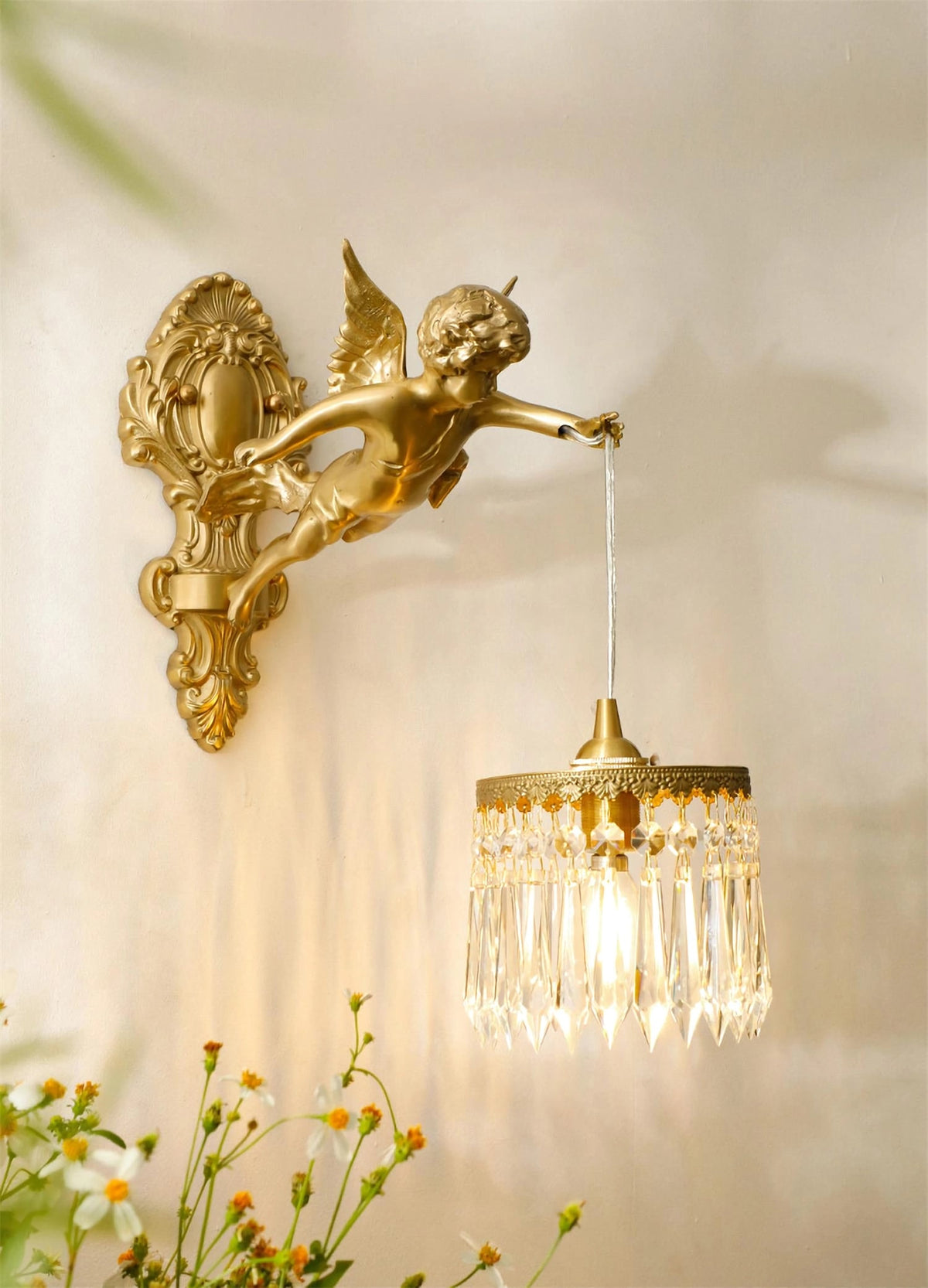 Above a spray of small daisies, a Clérachic cherub wall light lets its crystal-fringed shade glow warm on a cream wall.