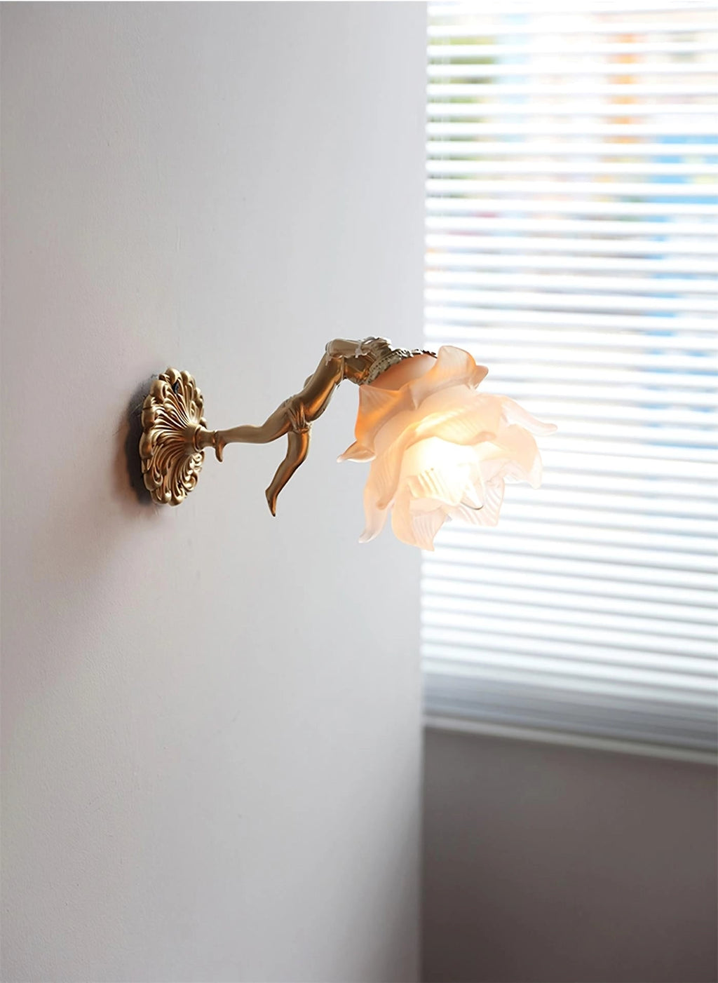 Seen from the side, a Clérachic cherub wall light with a rose glass shade projects from a grey wall beside a window with blinds.