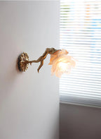 Seen from the side, a Clérachic cherub wall light with a rose glass shade projects from a grey wall beside a window with blinds.