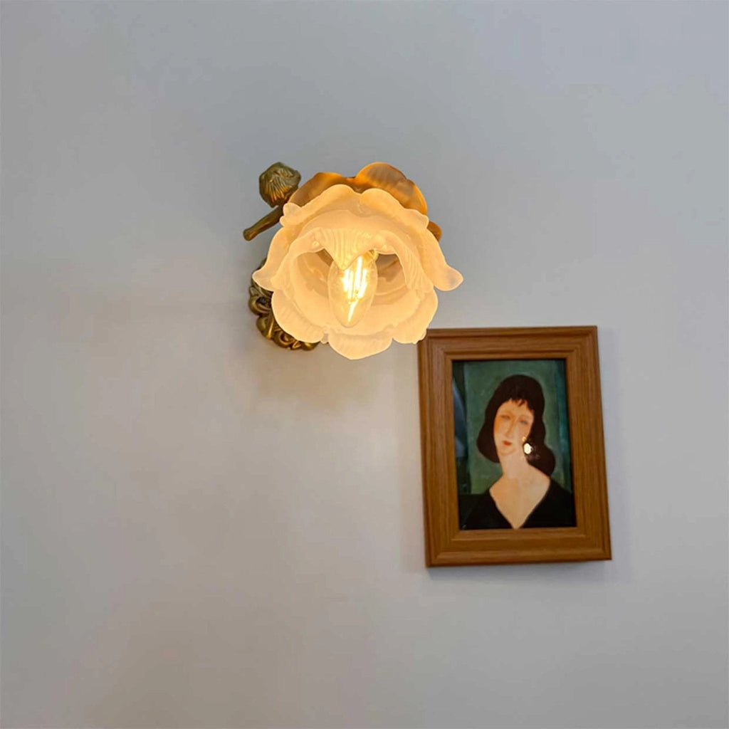 A lit Clérachic wall light with a rose-shaped glass shade hangs above a small framed portrait on a grey-blue wall.