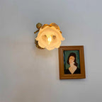 A lit Clérachic wall light with a rose-shaped glass shade hangs above a small framed portrait on a grey-blue wall.