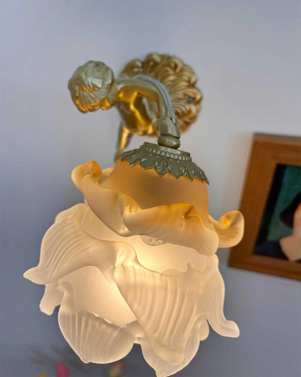 Looking up at the glass rose shade of this Clérachic wall light, with the cherub and a framed picture behind it.