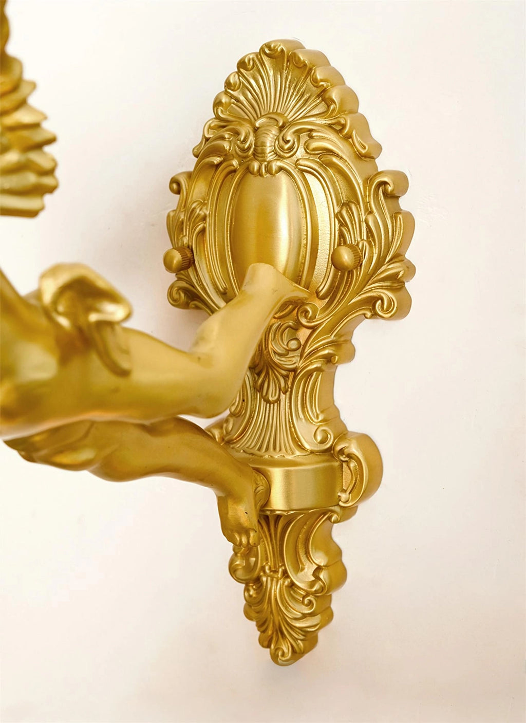 Close view of the scrolled, shell-topped back plate and the cherub's arm on this Clérachic wall light, in a gold-toned finish.