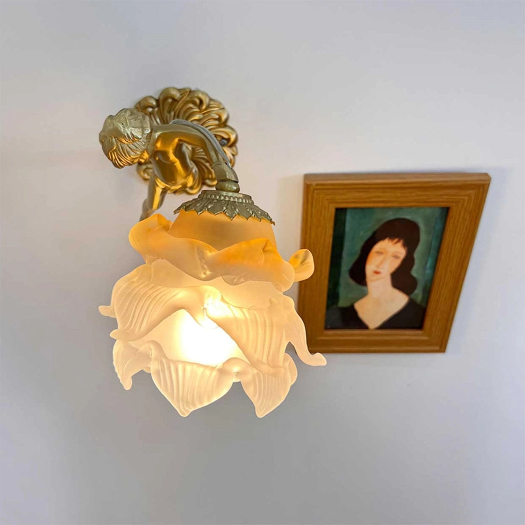 A Clérachic cherub wall light with its frosted rose shade lit warm, hung close to a framed portrait on a blue-grey wall.