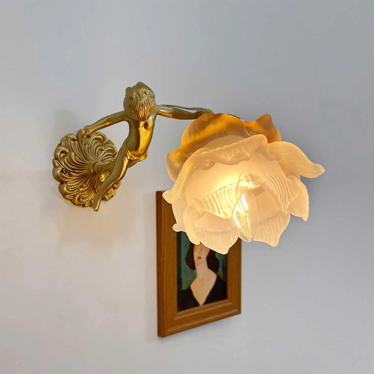 Side-on, a Clérachic cherub wall light with a lit rose shade projects from its round back plate above a framed portrait.
