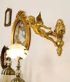 Beside a round gilt frame and a table lamp, a Clérachic cherub wall light hangs its frosted rose shade over a cream wall.
