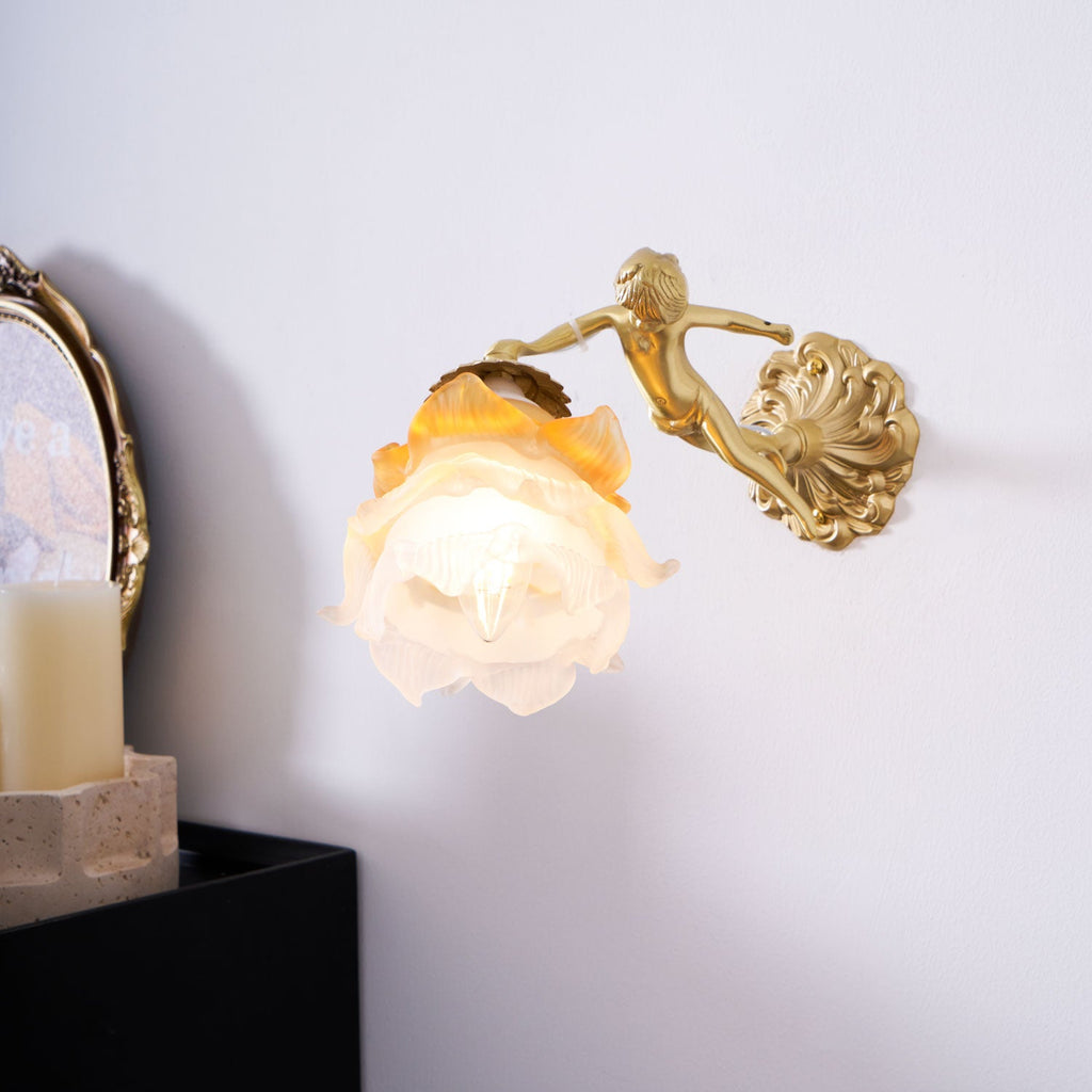 A Clérachic cherub wall light with a lit rose shade juts from a white wall beside a pillar candle and a round framed picture.