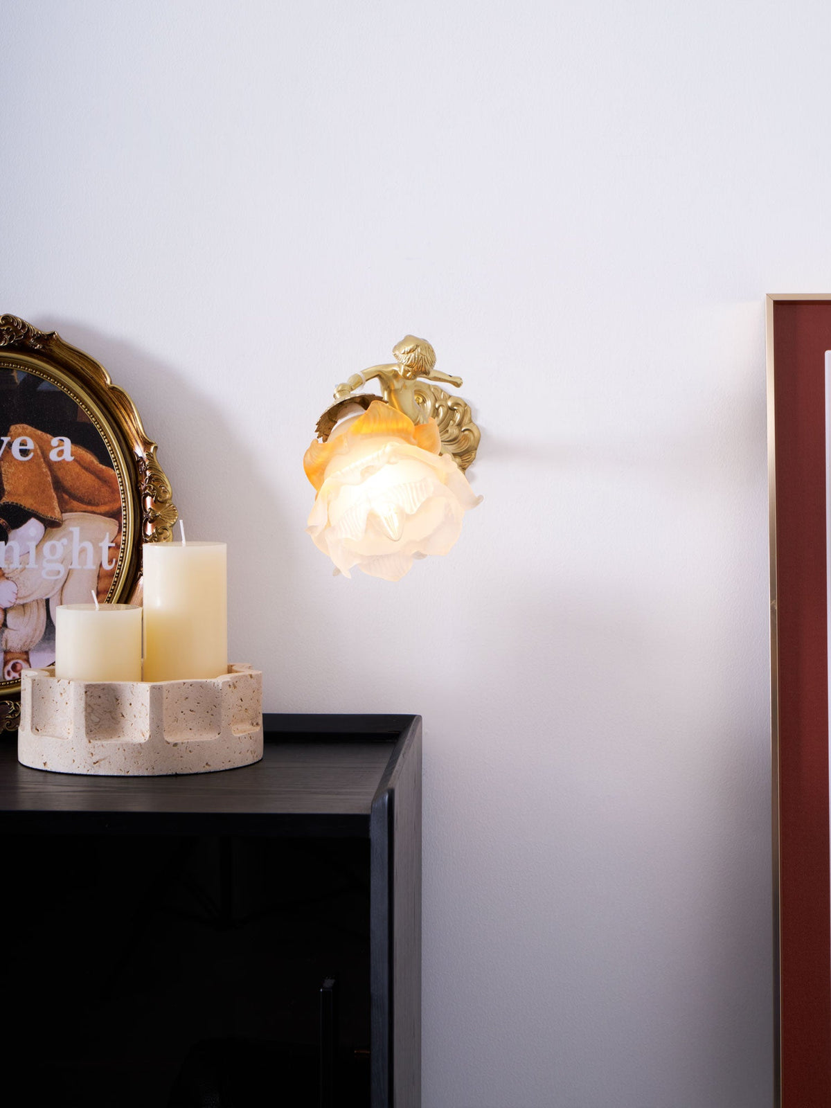 At a distance, a Clérachic cherub wall light with a rose shade sits on a white wall above a black cabinet and two candles.