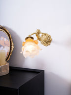 A Clérachic wall light with a frosted rose shade hangs on a plain white wall, with a candle and framed picture to the left.
