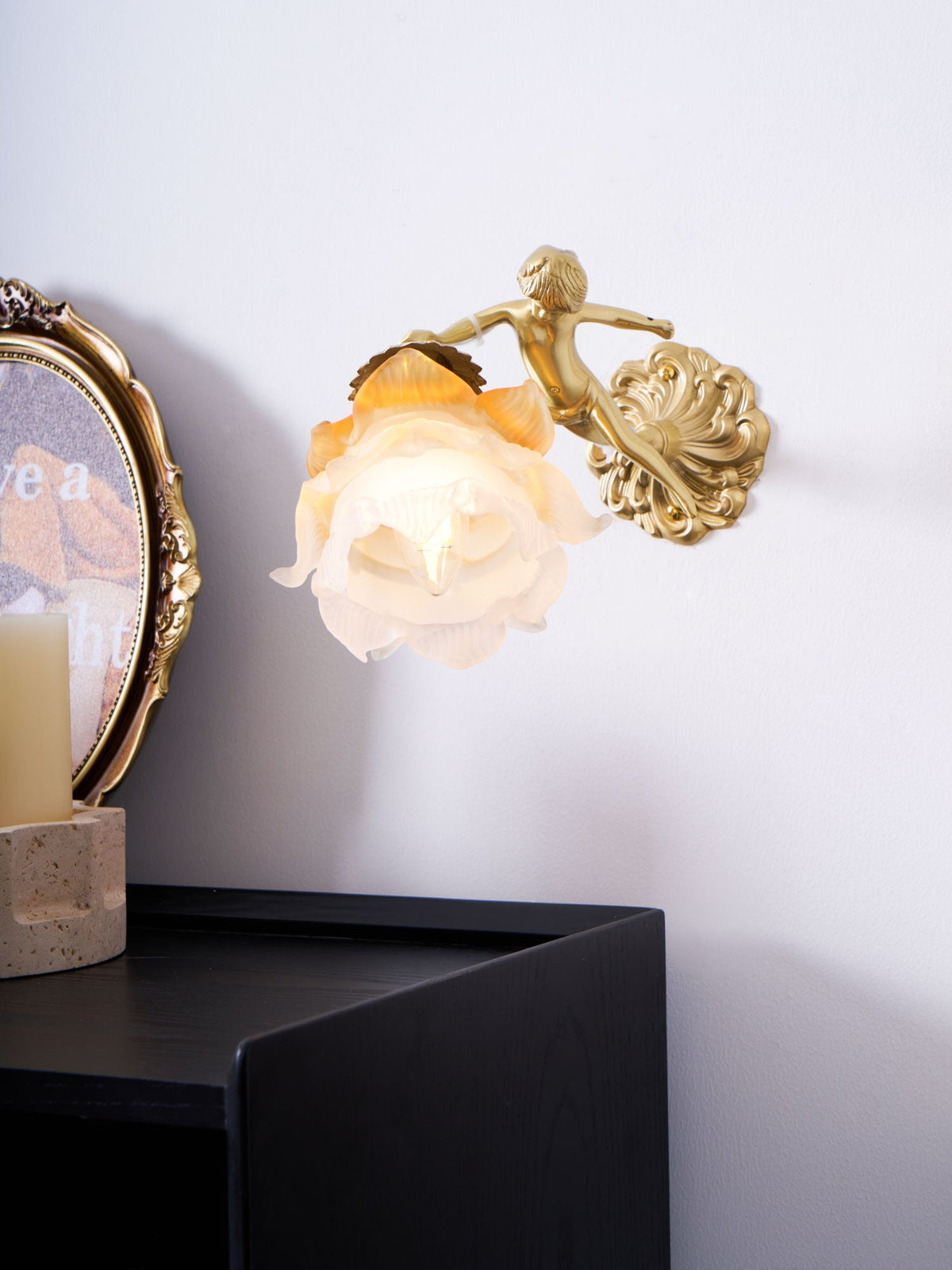 The small cherub and round back plate of this Clérachic wall light, with its rose shade lit above a black cabinet.