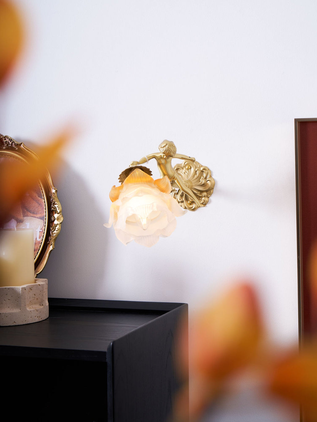 Through soft autumn-leaf blur, a Clérachic cherub wall light glows on a white wall above a black cabinet.