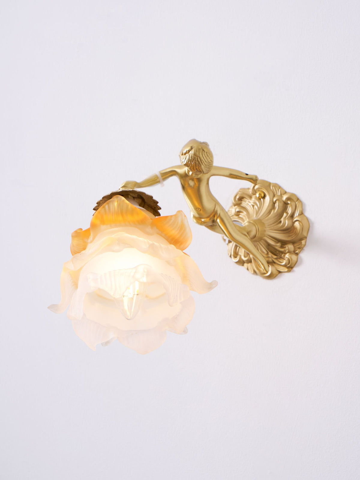A Clérachic cherub wall light with a lit rose shade and a scrolled round back plate, on a clean white wall.