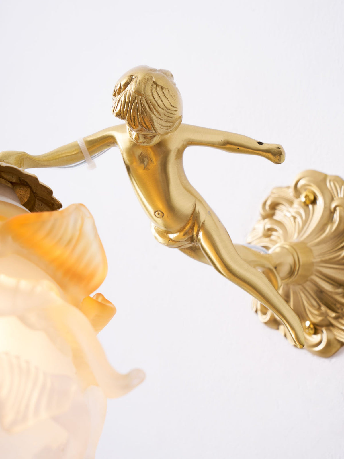 Close view of the cherub, a gold-toned scroll back plate and the edge of the glass shade on this Clérachic wall light.