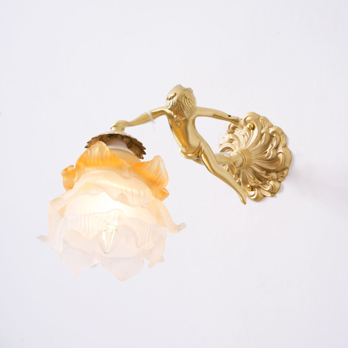 A Clérachic cherub wall light with an amber-tipped frosted rose shade, seen from slightly below against a white wall.