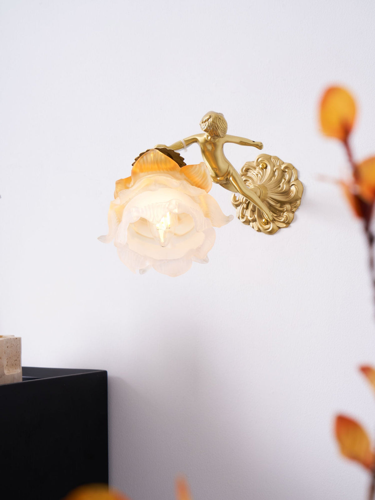 A Clérachic cherub wall light with a rose shade, small against a white wall with orange autumn leaves in the foreground.