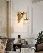 A Clérachic cherub wall light on a panelled white wall above a dark side table with picture frames and a green plant.